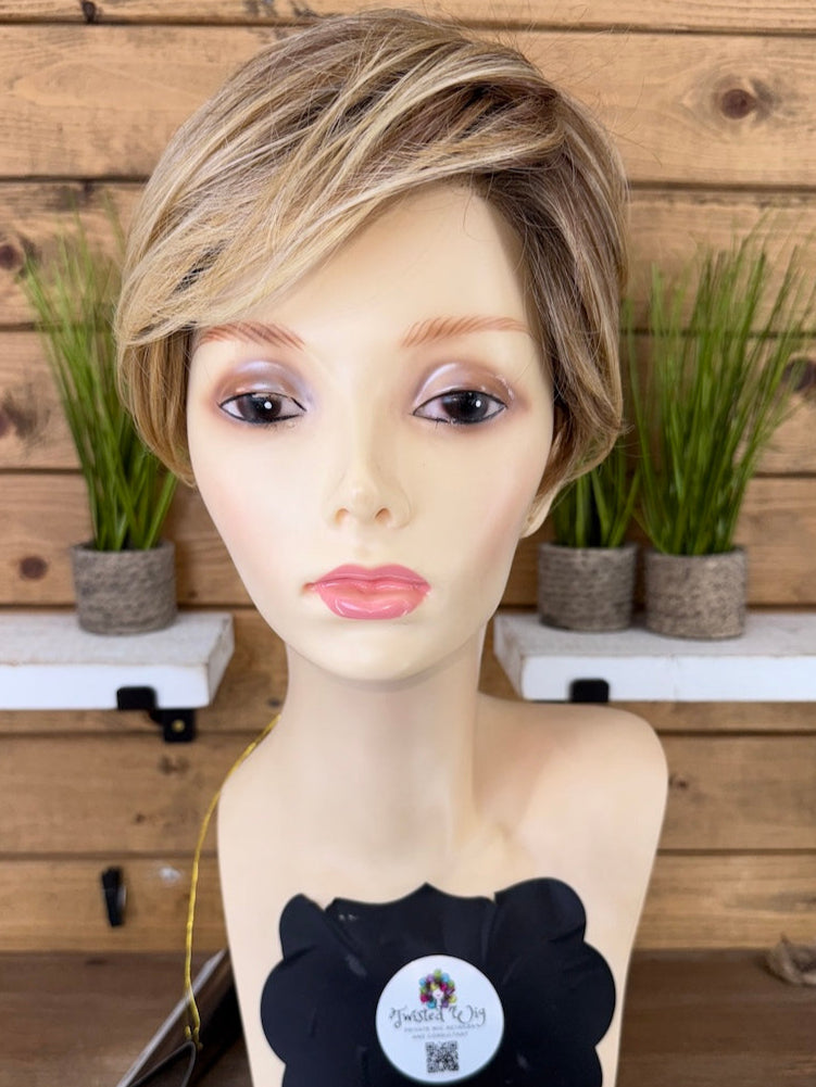 Mannequin head with a wig and black bow on a wooden surface with a wooden wall and plants in the background.